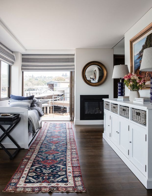 Bright, refined living room with dark timber flooring, a long patterned runner rug, a white built-in storage cabinet with woven basket drawers, and a black fireplace. A sofa sits beside large windows and a balcony door fitted with striped Roman blinds, while framed artwork, a round mirror, lamps, and floral decor add traditional accents. The room opens to an outdoor balcony with a leafy suburban outlook.