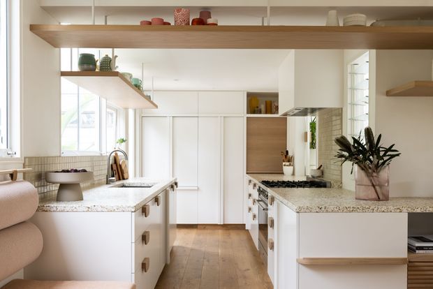 Bright contemporary kitchen with white cabinetry, a central island, pale stone benchtops, warm timber flooring and open timber display shelving. Large windows provide abundant natural light, while integrated appliances and a tiled backsplash create a clean, refined fit-out.
