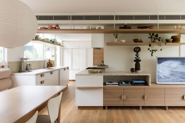 Bright open-plan kitchen and living area with warm timber flooring, white cabinetry, a stone kitchen island, integrated shelving, and a long timber entertainment unit. A dining table occupies the foreground, while large windows and layered open shelving create an airy, contemporary feel.
