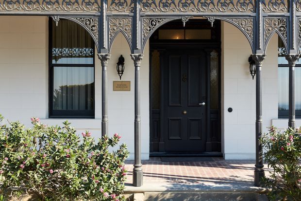Front entrance of a heritage-style building with an ornate cast-iron verandah, decorative arches and columns, a black paneled timber door, white horizontal wall cladding, and a patterned brick-paved entry. Established shrubs line the foreground.