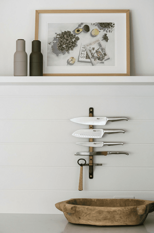 A carefully styled kitchen vignette with a white shaker-style cabinet or wall panel backdrop. A vertical magnetic knife rack holds several chef's knives and a serrated knife, with a rustic wooden-handled utensil below. On the upper ledge are two matte ceramic bottles and a framed still-life print in a natural timber frame. A large rough-hewn wooden bowl sits on the countertop in the foreground. The palette is soft white, natural timber, charcoal, and muted olive tones, creating a calm contemporary rustic atmosphere.