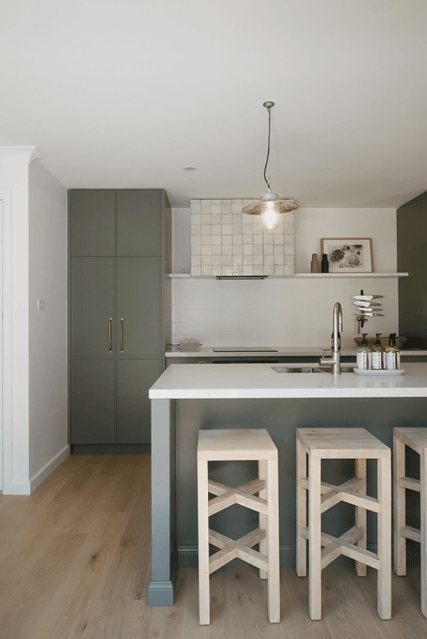 A compact, contemporary kitchen with a large white island and integrated sink, grey-green full-height cabinetry, pale timber flooring, and a tiled splashback. Three light timber bar stools sit beneath the island overhang. A simple pendant light, open shelf, and small decorative objects complete the calm, muted interior.