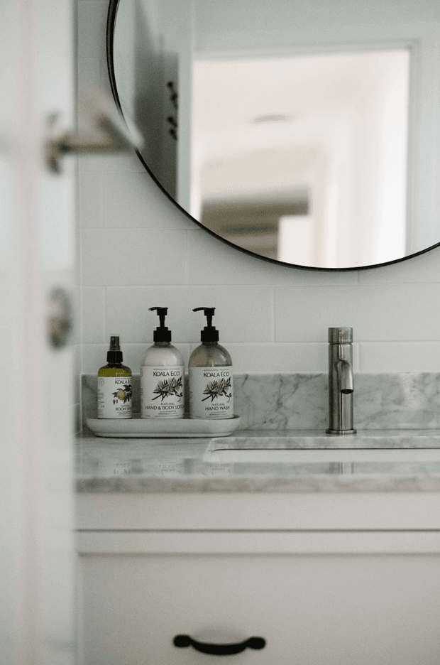 Close-up view of a refined bathroom vanity with a white inset basin, chrome mixer tap, pale marble-look benchtop and splashback, white tiled wall, white cabinetry, and a large round black-framed mirror. Neutral styling and natural personal-care products sit on a small tray beside the basin.