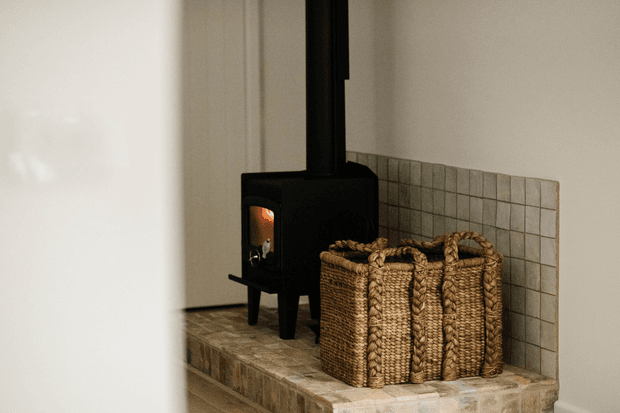 A compact black freestanding wood-burning stove with a visible fire sits on a raised tiled hearth in a minimalist interior corner. A tall black flue rises to the ceiling, while three woven natural-fibre baskets are arranged beside the stove against a tiled surround. The scene is softly lit with warm, earthy tones and simple pale walls.