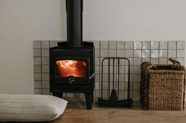 A compact black wood-burning stove with a visible fire sits against a light grey square-tile wall. A dark metal chimney rises vertically from the stove, with a matching fireguard and hearth tools beside it. A woven natural-fibre basket is positioned to the right, while a large off-white floor cushion occupies the foreground. The scene has a rustic, warm and cozy character with muted natural tones.