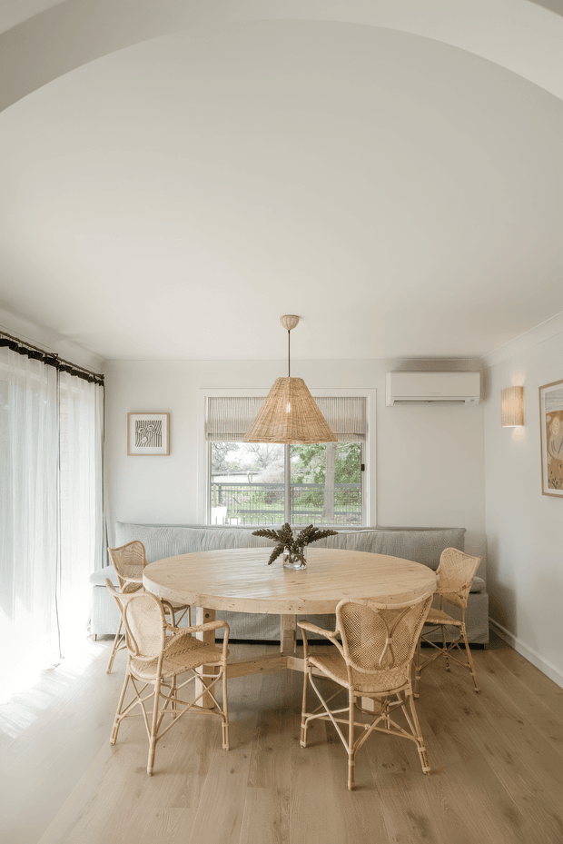 A bright, serene dining area with a large round light-wood dining table surrounded by woven rattan chairs. A matching woven pendant light hangs above, with a pale upholstered bench or sofa behind the table. Sheer curtains, a central window overlooking greenery, light timber flooring, soft white walls, framed artwork, and a wall-mounted split-system air conditioner create a warm, relaxed interior.