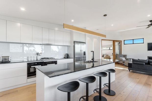 Bright open-plan contemporary kitchen with extensive white flat-panel cabinetry, integrated stainless-steel appliances, a large island with a dark stone benchtop and black bar stools. Light timber flooring continues into the adjacent living area, which includes a dark sofa and timber-framed glazed doors. A simple timber feature beam spans above the kitchen, and recessed ceiling lights provide even illumination.