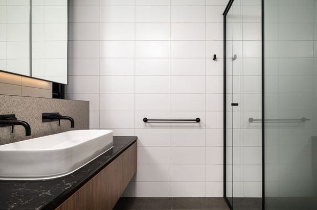 A contemporary bathroom with a floating timber-veneer vanity, dark stone benchtop, white integrated basin, black wall-mounted tapware, large white wall tiles, and a frameless glass shower enclosure with black hardware. A large mirror and recessed warm lighting add brightness and depth.