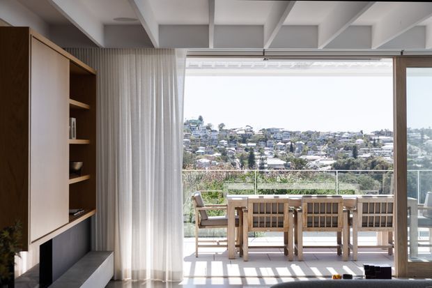 Bright contemporary interior opening onto a balcony or terrace through wide sliding glass doors. The room features built-in dark timber joinery with open shelving and cabinetry, a white exposed-beam ceiling, sheer curtains, and a timber outdoor dining setting overlooking a leafy suburban landscape.