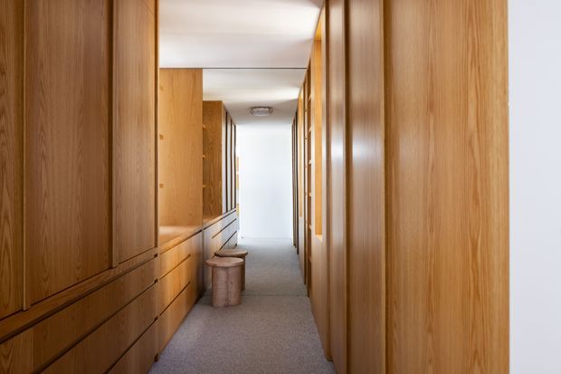A narrow, long hallway lined on both sides with extensive floor-to-ceiling timber cabinetry and panelled walls. The corridor has a light carpeted floor, white ceiling and walls, warm natural wood tones, recessed storage, and a small stool or ottoman along the left side. Bright daylight draws the eye toward the far end.