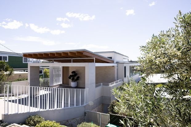Exterior view of a contemporary multi-level residence with white rendered and light brick-like wall surfaces, extensive white metal balustrades, and a prominent flat timber-lined pergola or canopy over the entry terrace. The home is set on a sloping site, with stepped outdoor levels and mature planting partially screening the facade. Bright, clear daylight emphasizes the clean geometric composition.