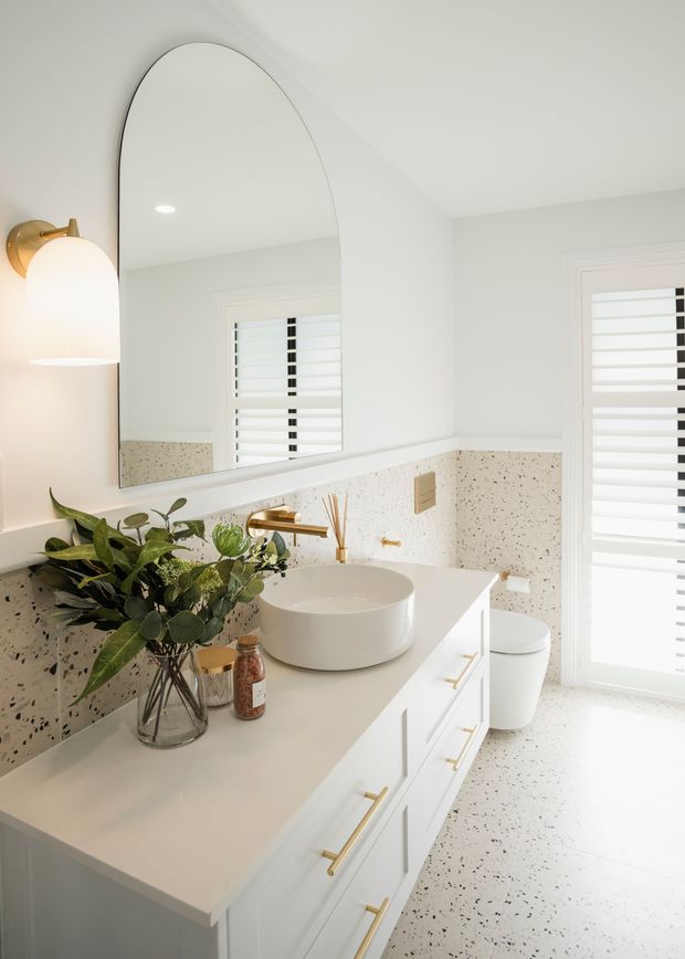 Bright, contemporary bathroom featuring a long white vanity with integrated storage, a round vessel basin, brushed brass tapware and handles, an arched wall mirror, and white terrazzo-style wall and floor finishes with dark aggregate flecks. White plantation shutters and a toilet are visible, along with a leafy arrangement on the vanity.