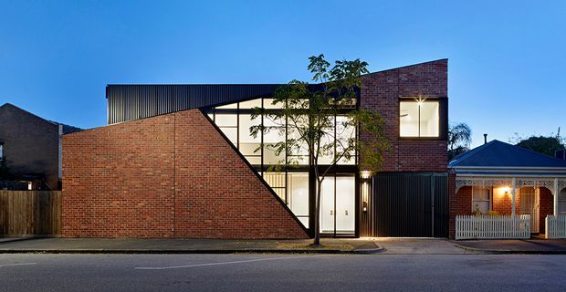 Street-facing view of a contemporary multi-storey urban residence at dusk. The building features a strong geometric composition with exposed red brick walls, black vertically ribbed metal cladding, extensive illuminated glazing, and a sharply angled brick facade forming a dramatic triangular profile. A traditional neighbouring cottage is visible to the right.