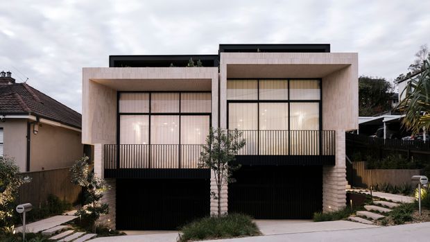 Front exterior view of a contemporary two-storey residential building with two projecting box-like upper volumes, pale textured masonry or concrete façades, extensive floor-to-ceiling glazing, black metal-framed windows, and black vertical batten screening at the balconies and lower level. The home sits on a suburban street with stepped pedestrian paths, landscaped planting, and neighbouring houses visible at the sides.