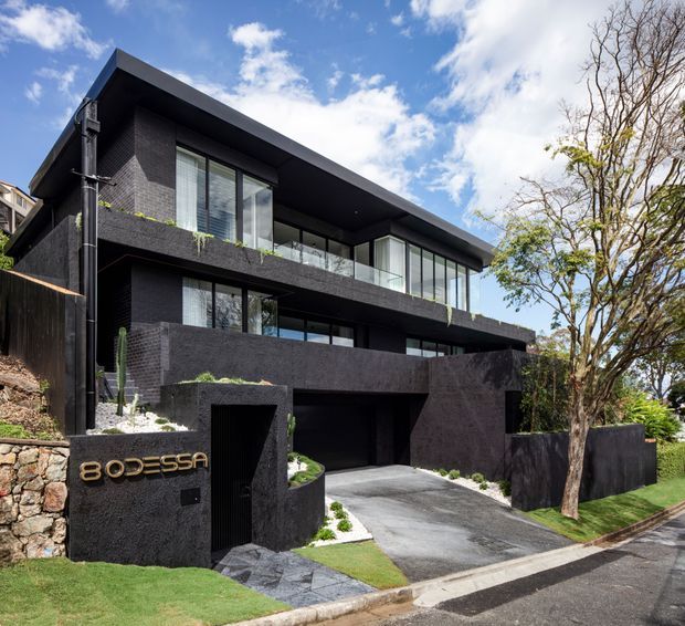 Street-level exterior photograph of a striking multi-storey contemporary residence with dark brick or masonry walls, broad projecting flat roof planes, extensive floor-to-ceiling glazing, balconies with glass balustrades, and an integrated driveway leading to a concealed garage. The home is set on a sloping suburban site with landscaped planting and mature trees.