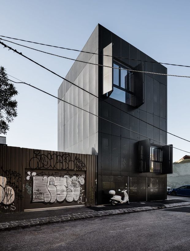 Street-level exterior view of a narrow, multi-storey contemporary building clad in dark perforated metal screening. The angular upper facade and projecting glazed windows create a strong geometric form, with a lighter perforated side wall and a graffiti-covered garage frontage at street level. Overhead utility wires and parked vehicles are visible in the urban streetscape.