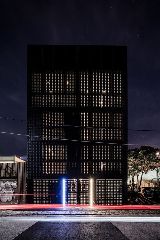 Night-time street view of a tall, narrow contemporary building with a dark perforated metal facade. Warm interior lights glow through the patterned screen across multiple levels, while bright blue and warm-white entrance lighting highlights the ground floor. Urban streetscape, adjacent graffiti-covered structure, trees, overhead wires, and wet pavement are visible.