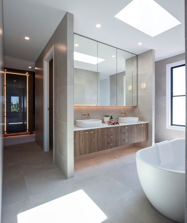 Bright contemporary bathroom with a double floating timber vanity, two vessel basins, brushed gold tapware, large mirrored cabinetry, pale grey tiled walls and floor, a freestanding white bathtub, skylights, and a dark-framed window. A darker adjoining corridor or dressing area is visible beyond the bathroom entry.