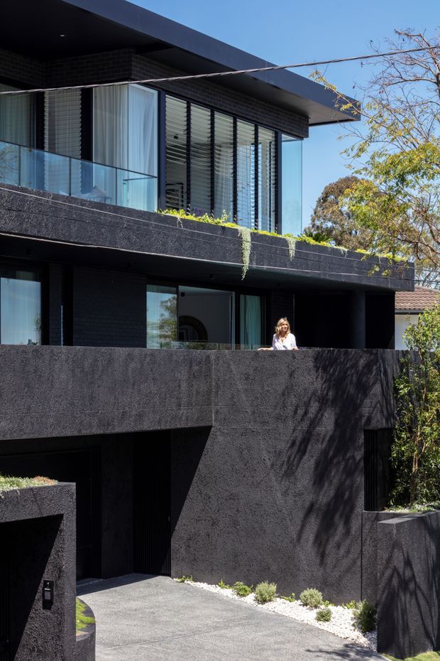 Exterior view of a contemporary multi-storey residential building with a dramatic black textured facade, broad flat roof overhangs, deep recessed openings, extensive glazing, external blinds, glass balcony balustrades, and layered terraces. A paved driveway leads toward a recessed garage or entry, with minimal planted borders and mature trees around the property.