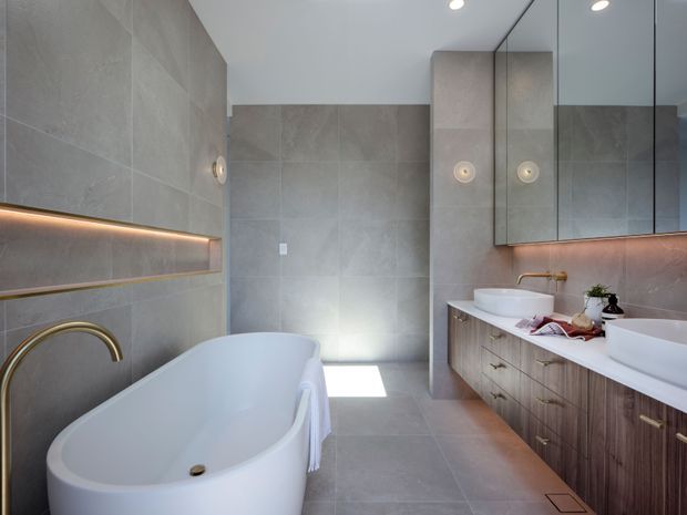 Contemporary bathroom with large-format grey stone-look wall and floor tiles, a freestanding white bathtub, twin white vessel basins, timber-look vanity cabinetry, mirrored wall cabinets, and warm integrated strip lighting. The space has a clean, refined appearance with brass tapware and fittings.