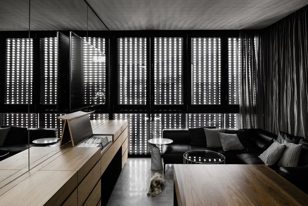 A dramatic contemporary living room and media space with a black leather sectional, round glass coffee table, wood cabinetry and a large wood-topped island or console. Mirrored walls reflect the room, while patterned perforated window screens create strong dotted light across the dark interior. The ceiling and walls feature dark textured finishes, producing a high-contrast, cinematic atmosphere.