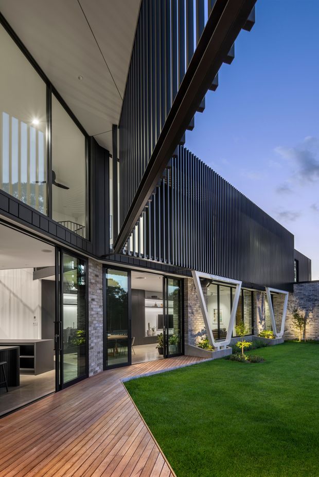 Exterior architectural photograph of a contemporary multi-level residence viewed from a landscaped courtyard. The building combines dark vertical metal screening and cladding, extensive black-framed glazing, exposed stone masonry, projecting timber beams, and distinctive angled structural supports. A timber deck leads through glazed doors to a neatly maintained lawn and planted garden, while interior living spaces are visible through the windows.