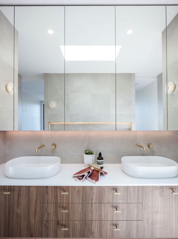 Contemporary double-vanity bathroom with two white vessel basins on a white countertop, brushed brass wall-mounted tapware, large mirrored cabinetry, pale stone-look wall finishes, integrated warm lighting, and a dark timber vanity with brass pulls.