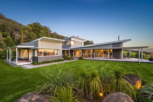 Exterior architectural photograph of a contemporary single-storey home set within a landscaped, sloping rural or semi-rural site. The house has articulated low-pitched roof forms, light vertical metal cladding, dark masonry walls, extensive glazing, covered outdoor living areas, and a long slim carport-like canopy. Native grasses, rocks, lawn, and mature bush vegetation surround the building.