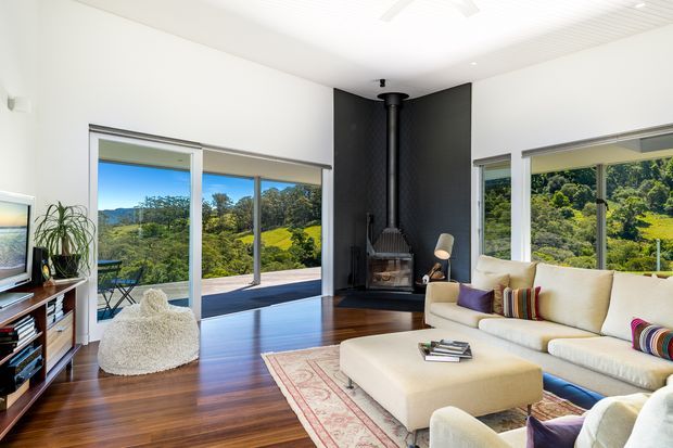 Bright contemporary living room with expansive glazing overlooking lush wooded hills. The room features dark timber flooring, a white sectional sofa and ottoman, a patterned area rug, a black feature fireplace wall with a freestanding wood-burning stove, and sliding glass doors opening to an outdoor deck. A low white slatted ceiling and clean white walls create an airy interior.