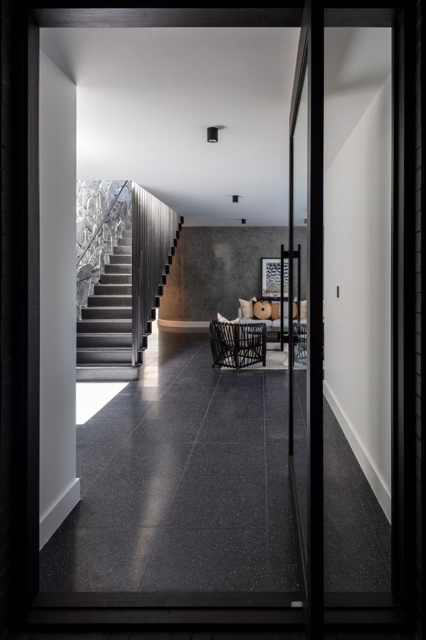 A dark, refined residential entry hall viewed through a black-framed glazed doorway. The space features large dark stone or porcelain floor tiles, white walls and ceiling, a sculptural open staircase with dark treads and vertical black balustrade, a textured grey feature wall, and a partially visible contemporary seating area. Natural light enters beside the staircase, creating strong contrast against the moody interior.