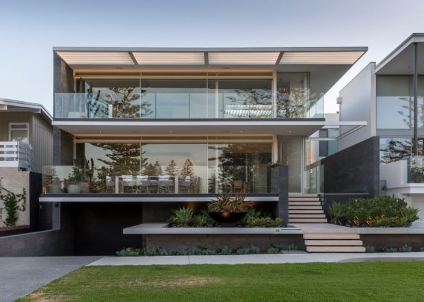 Front exterior view of a contemporary multi-level residence with extensive floor-to-ceiling glazing, projecting flat roof planes, broad horizontal balconies, dark and light facade surfaces, and landscaped frontage. A covered upper terrace, outdoor dining area, stairs, and integrated planter beds are visible.