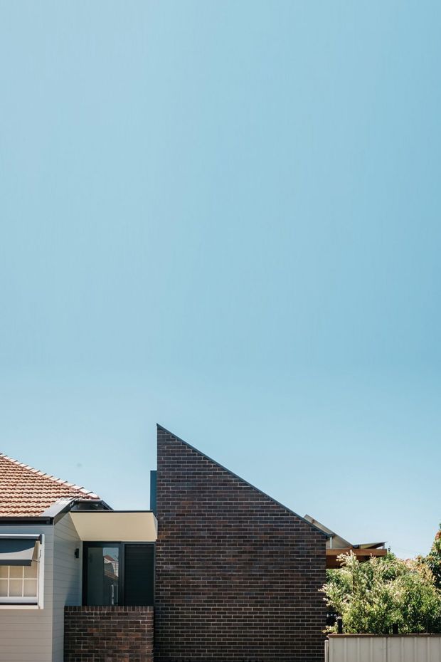 Exterior architectural photograph of a contemporary single-storey residential extension or house, featuring a prominent triangular dark brick gable wall, light horizontal cladding, dark-framed glazing, and a tiled roof section. The building is viewed from outdoors against a clear blue sky, with neighbouring roof forms and garden elements at the edges.