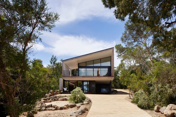 A modern two-storey house set within a wooded landscape, viewed from the front. The building has a strongly geometric form with a single-sloping roof, broad upper-level glazing, a covered balcony, dark lower-level openings, and an integrated carport. A pale driveway curves through naturalistic rock landscaping and native-looking vegetation toward the house.