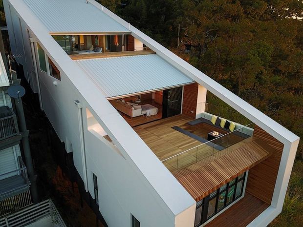 Aerial view of a narrow contemporary multi-storey residence with a distinctive white pitched roof and large open roof terraces. The upper terrace includes timber decking, outdoor seating, a glass balustrade, and a built-in square fire feature. Large glazed openings reveal a warm-toned interior, while surrounding mature trees frame the property.
