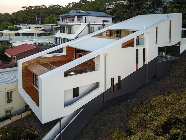 Aerial view of a contemporary multi-level residential house on a steeply sloping suburban site. The home has white rendered walls, bold angular roof planes, dark lower-level cladding, warm timber-lined balconies and recessed terraces, extensive glazing, and an elevated driveway approach. Other houses and dense vegetation surround the property.