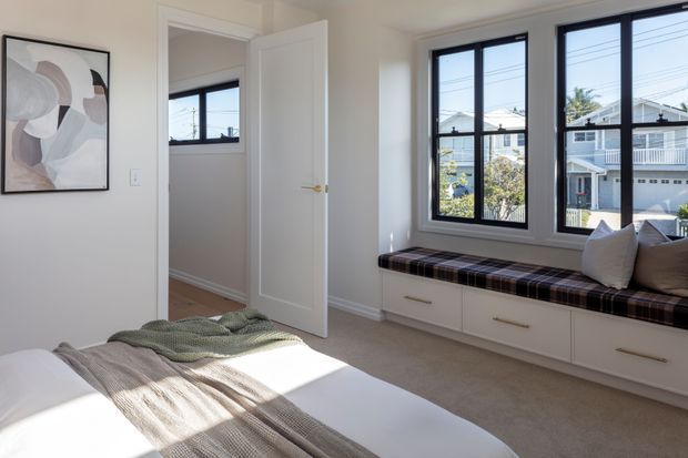 Bright, softly styled bedroom with a white upholstered bed, neutral bedding, a muted abstract artwork, and a built-in window seat with drawers beneath large black-framed windows. The room has light walls, carpeted flooring, and views of neighbouring homes through the glazing.