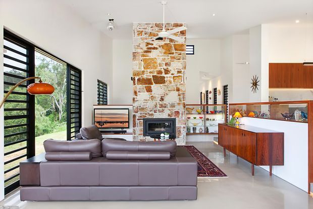 Bright open-plan living area with a large modular leather sofa facing a television and a central stone-clad fireplace chimney. The room includes extensive black-framed louvred glazing, timber mid-century-style cabinetry, a white kitchen island, open shelving, polished light flooring, and a vintage orange pendant lamp. The space has a clean contemporary shell with warm natural stone and retro-inspired furnishings.