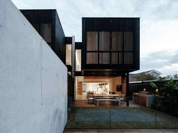Contemporary multi-level residential exterior viewed from a landscaped rear or side courtyard. The building features dark box-like volumes with vertical timber screening, a projecting upper-level section, warm timber soffits, and large open glazing at ground level. A paved outdoor dining area and planted boundary sit in front of the partially visible interior kitchen and living space, while a tall white rendered boundary wall frames the left side.