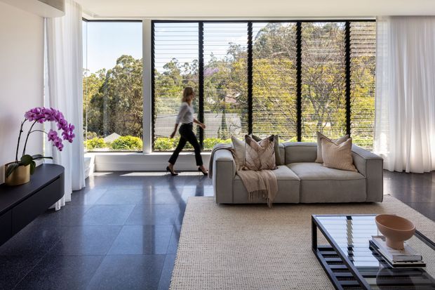 Contemporary living room with a grey sofa, neutral textured cushions and throw, large floor-to-ceiling windows with dark horizontal louvres, sheer curtains, glossy dark tiled flooring, a pale woven rug, and a black glass coffee table. The room is bright and visually connected to leafy surroundings.