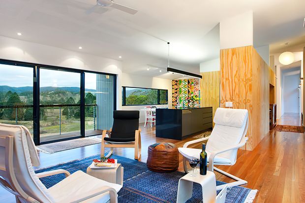Bright open-plan contemporary living area with expansive black-framed glazing overlooking a green landscape. The room features polished timber flooring, a large blue patterned rug, white lounge chairs, a black sideboard, a central island or kitchen zone, colourful wall artwork, and warm timber wall panelling. A hallway and additional interior spaces are visible to the right.