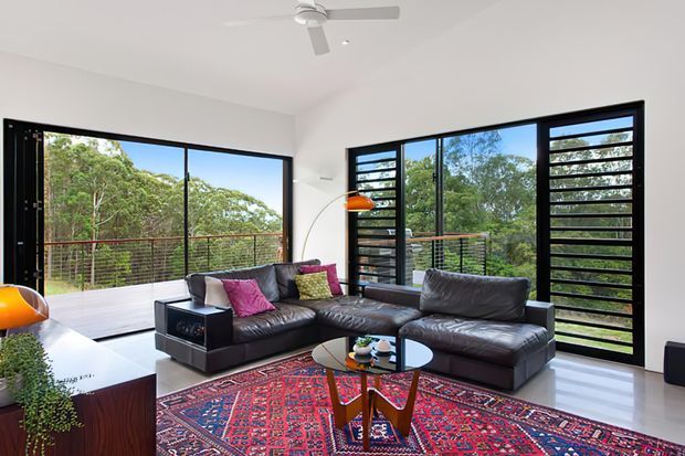 Bright contemporary living room with large black-framed glazed doors and louvred windows opening toward a leafy outlook and balcony. The room features two dark leather sofas, a patterned red rug, a round glass coffee table, orange accent lighting, white walls, polished light flooring, and a ceiling fan.