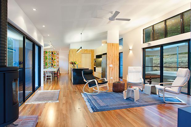 Bright open-plan modern living room with polished timber flooring, extensive black-framed glazing, a lounge seating area, ceiling fans, and a timber-clad structural column. The space includes a kitchen area toward the rear and a colourful decorative wall panel, with abundant natural light and a relaxed mid-century-influenced furnishing scheme.