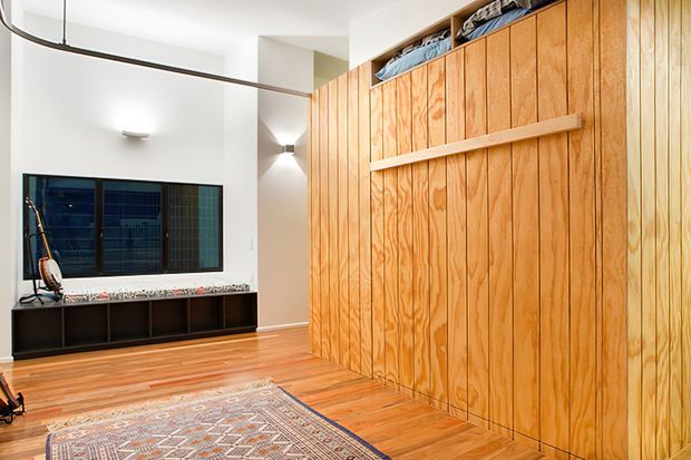 Bright contemporary interior featuring a full-height timber storage wall with vertical panelled doors and open shelving above. The room has warm timber flooring, a low dark built-in media console, a large window, curved black ceiling detailing, and a patterned area rug.