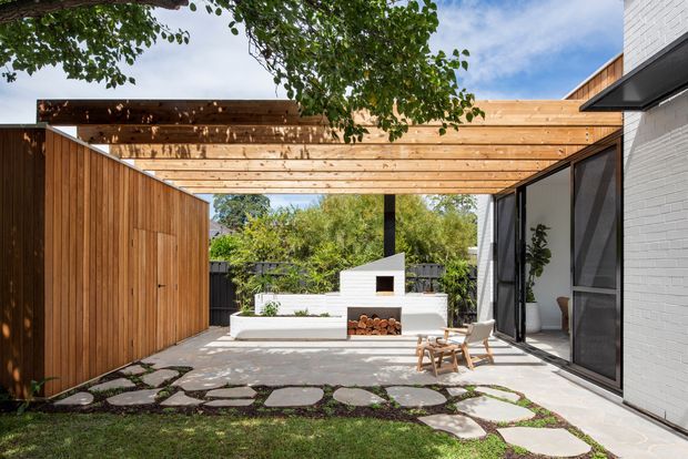 A landscaped courtyard patio framed by a timber-clad wall and a broad exposed timber pergola. The space features a white rendered outdoor fireplace with integrated log storage, concrete paving with irregular stepping stones, dark-framed glazing, and mature greenery. The overall design is contemporary and warm, with a strong indoor-outdoor connection.