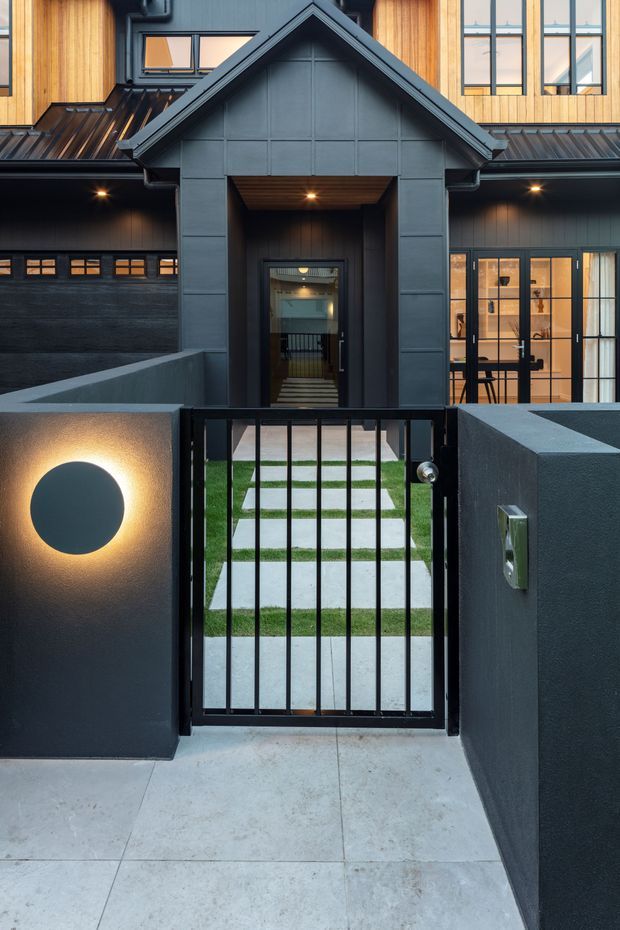 Front exterior view of a contemporary multi-level residence, centred on a covered gabled entry porch behind a black metal pedestrian gate. The facade combines dark painted cladding and exposed warm timber cladding, with large glazed doors and windows. A concrete-paved approach and narrow lawn with stepping stones lead to the entrance, while integrated wall lighting and a dark garage door reinforce the modern, moody presentation.