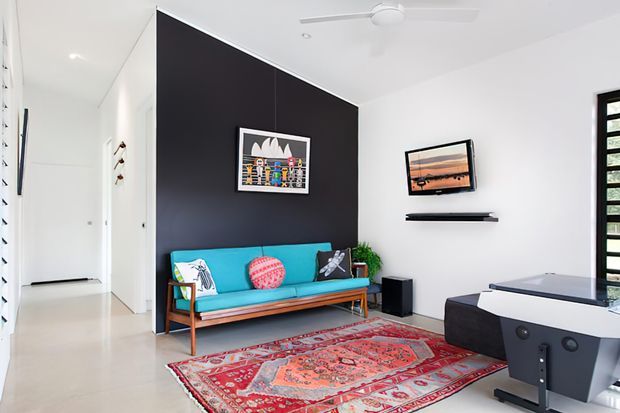 Bright, compact contemporary living room with a vivid turquoise mid-century-style sofa, patterned cushions, a large red Persian-style rug, black feature wall, wall-mounted artwork and television, and a partial foosball table. White walls, polished pale flooring, ceiling fan, and strong natural light create a clean, playful interior.