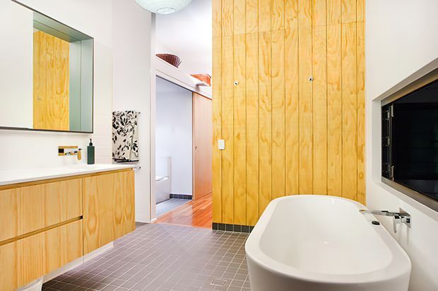 Bright contemporary bathroom featuring a freestanding white bathtub, timber wall panelling and matching timber vanity cabinetry, grey tiled flooring, a large mirror, and a wall-mounted television. An open doorway leads toward an adjacent area.