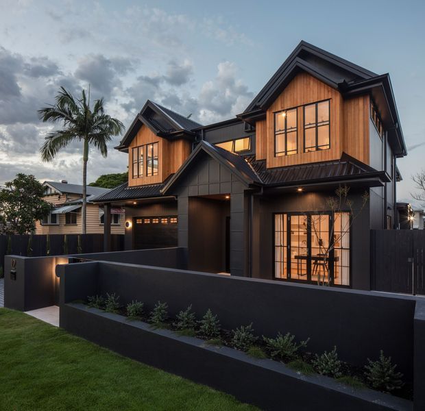 Exterior view of a contemporary multi-storey residence with steep gabled roof forms, dark cladding and trim, warm timber-clad upper-level volumes, and large black-framed windows. A low dark boundary wall and landscaped planter define the front yard, with a palm tree and neighboring homes visible in the background.