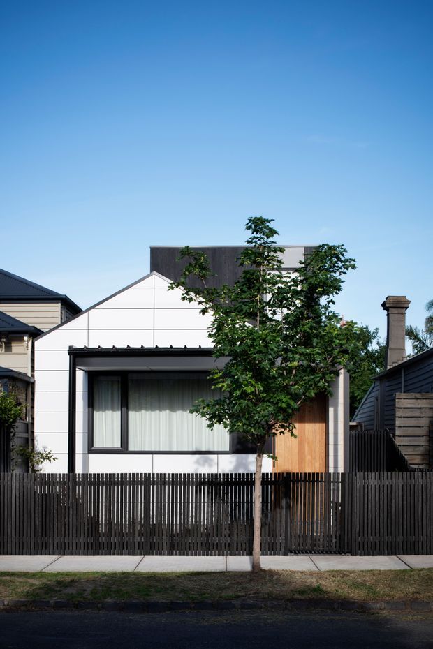 Street-facing view of a compact contemporary residence in an established suburban setting. The facade combines white panelled cladding with dark charcoal elements, a steep gabled roof form, a large curtained window, and a warm timber entry door. A dark vertical-slat fence and mature street tree partially screen the house, while neighbouring homes and a prominent masonry chimney are visible at the sides.
