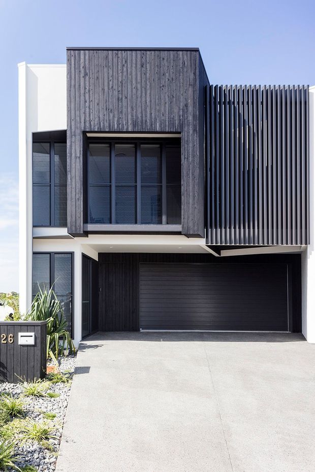 Front exterior view of a contemporary two-storey townhouse with a sharply articulated facade. The house combines dark vertical timber-look cladding, white rendered walls, expansive dark-framed glazing, and a prominent vertical slatted screen over the upper level. A wide dark garage door opens onto a pale concrete driveway, with a narrow landscaped strip of gravel and low planting along the frontage.